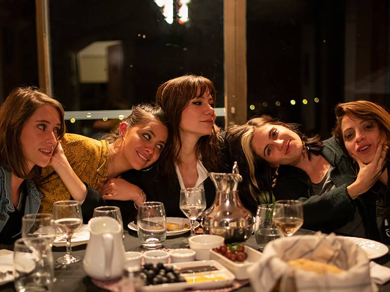 Fünf weiblich gelesene Personen sitzen an einem Tisch in einem Restaurant und scherzen miteinander. Hinter den Fenster ist es dunkel, auf dem Tisch stehen Weingläser und Weintrauben.